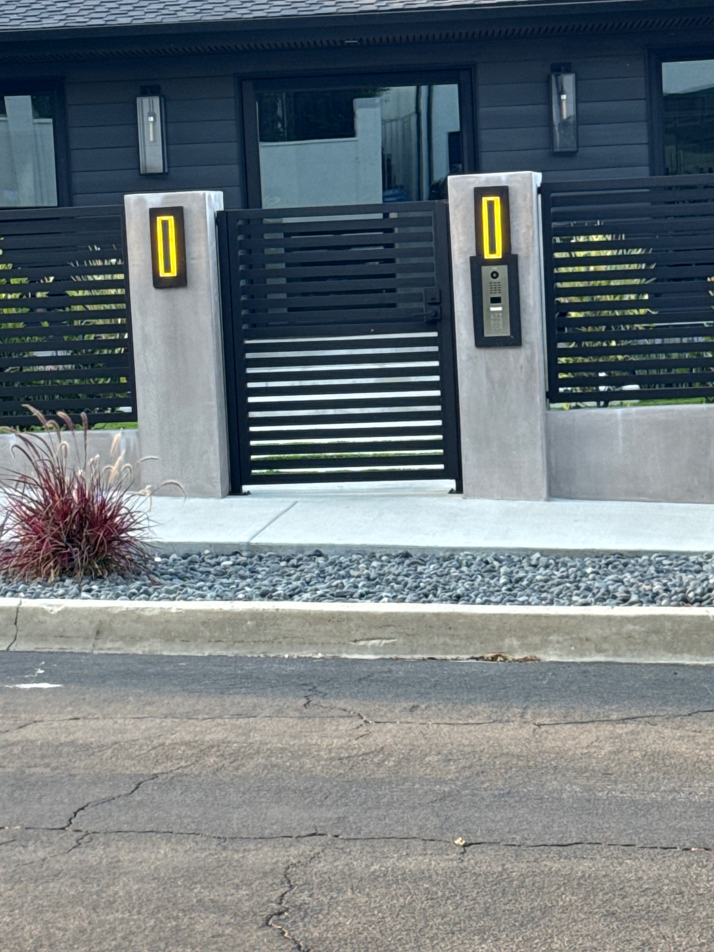 Black slat entry gate flanked by concrete pillars with yellow LED sconces and keypad intercom